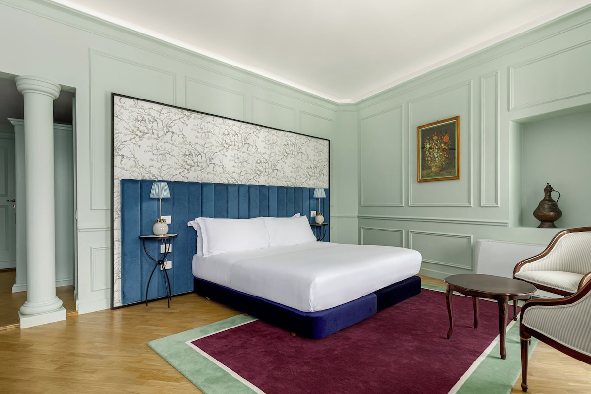 A bedroom with a large bed, a nightstand, a chair, and a painting on the wall.