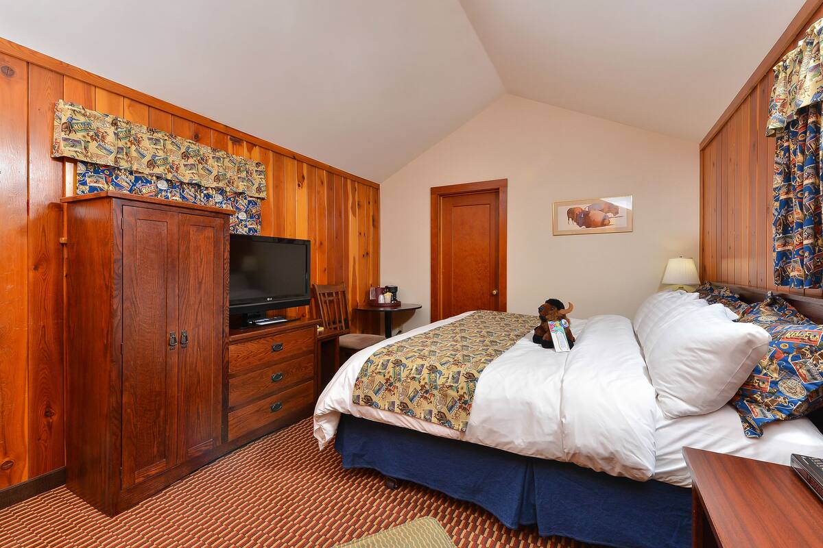 A bedroom with a wooden wardrobe, a television, a bed with patterned bedding, a small table, and a lamp.