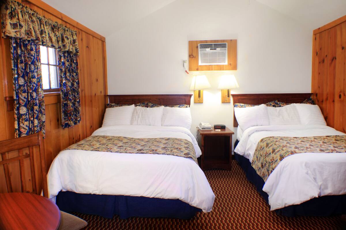 A hotel room with two beds, wooden headboards, a small table, and a window with curtains.