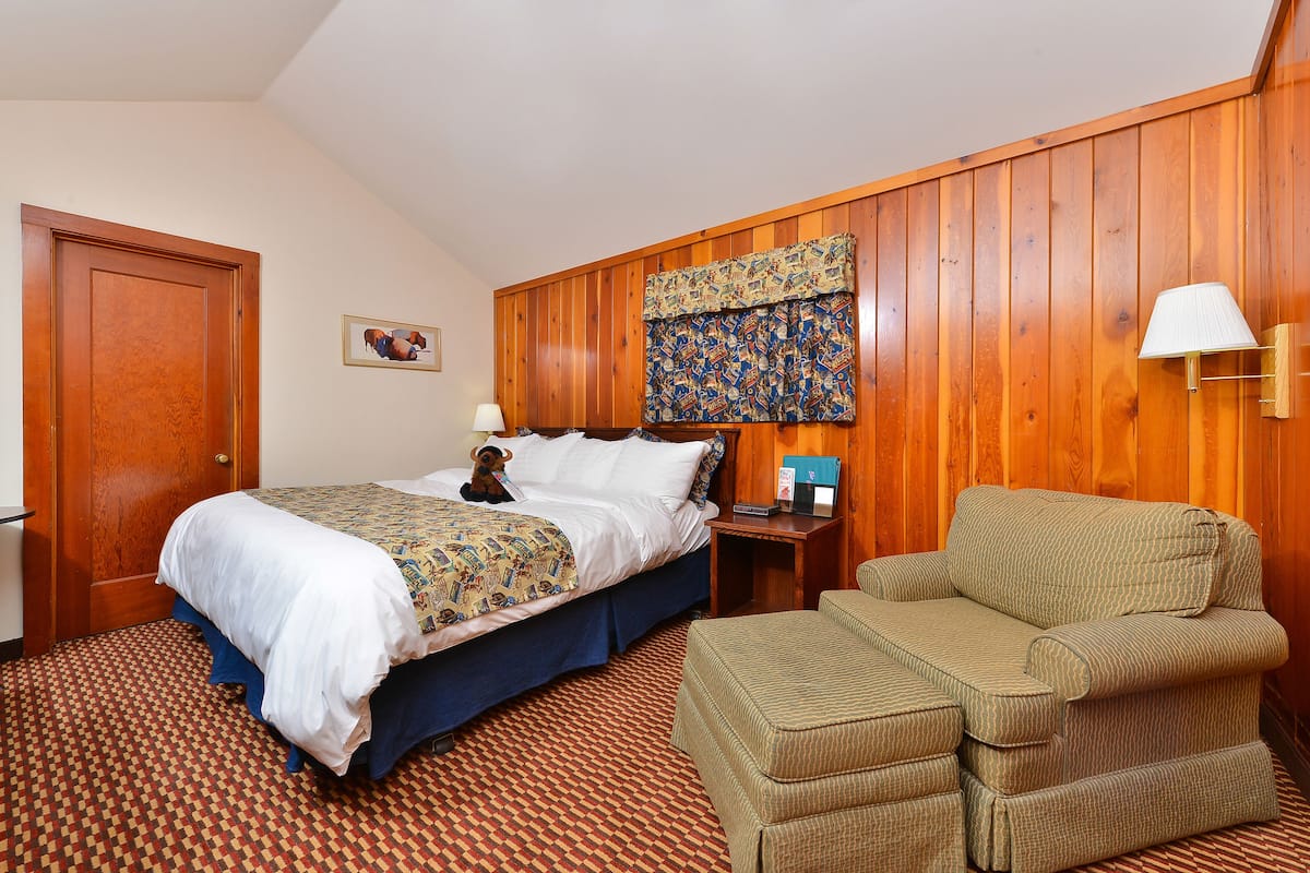 A hotel room with a wooden paneled wall, a bed with a patterned bedspread, a bedside table with a lamp, and a green upholstered chair.