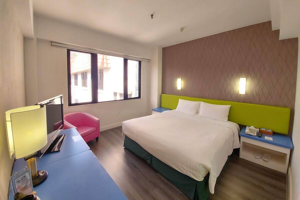 Habitación de hotel con una cama, un escritorio con lámpara, una silla, una mesita de noche con teléfono y control remoto, y una ventana con vistas a edificios.