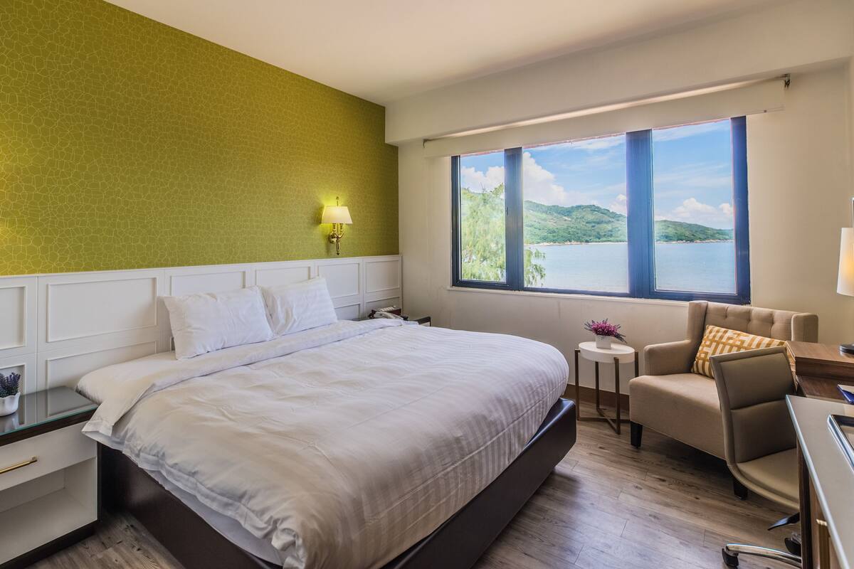 Habitación de hotel con una cama grande, un escritorio con una silla, una mesita de noche con una lámpara y una ventana con vistas al lago y las colinas.