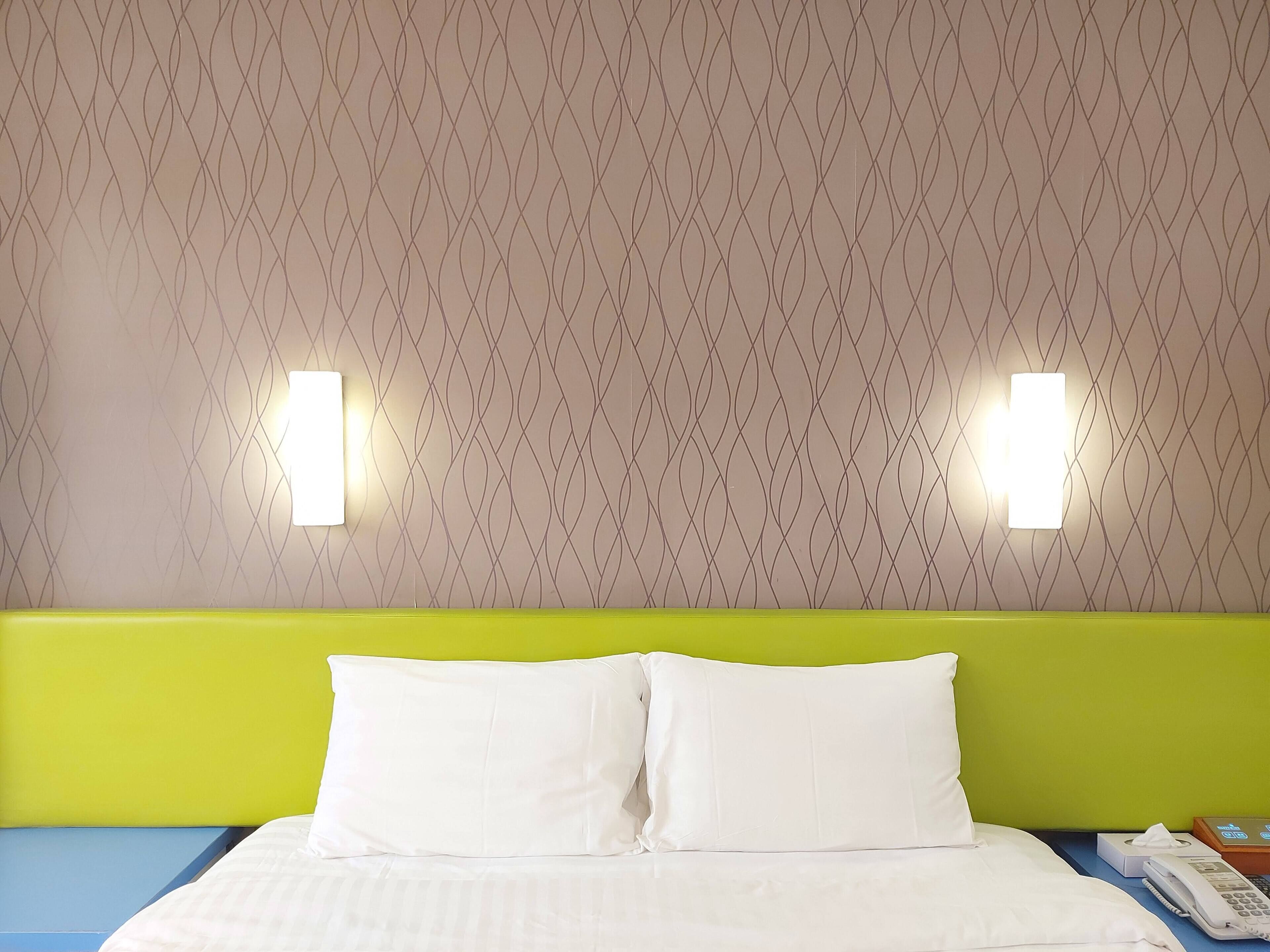 Camera d'albergo con carta da parati fantasia, una testiera verde, biancheria da letto bianca e un comodino con un telefono e una scatola di fazzoletti.