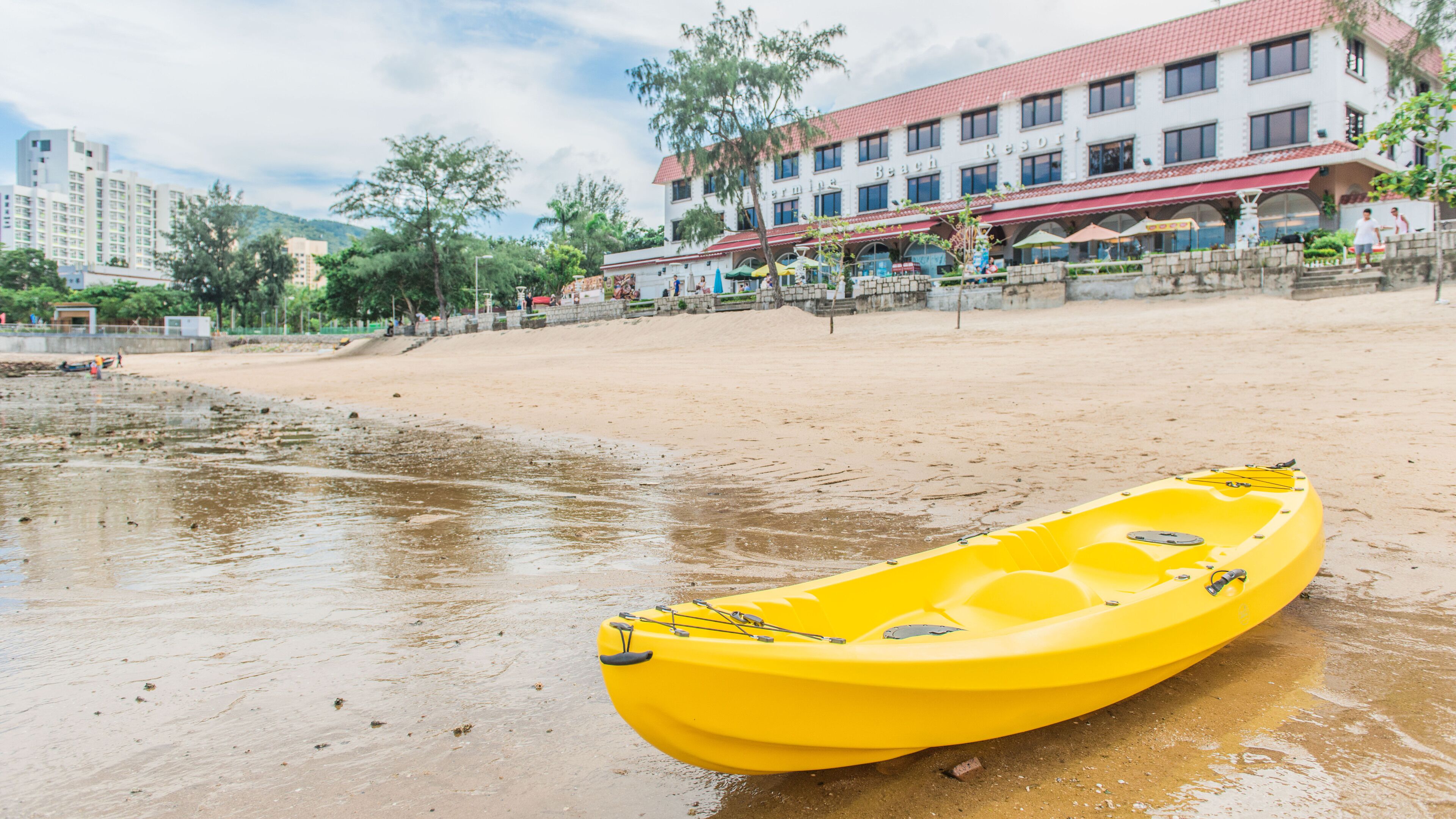 Un kayak amarillo sobre una playa arenosa, con un edificio que dice "Beach Resort" al fondo.