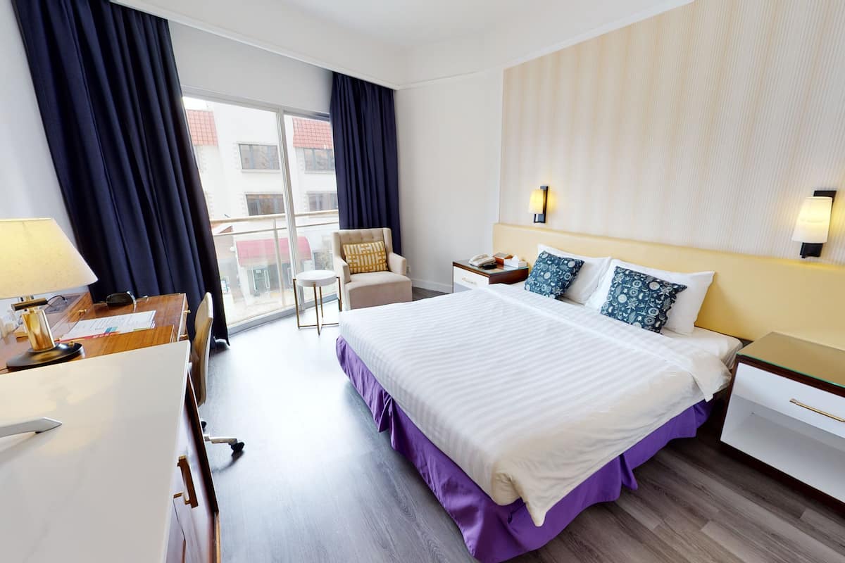 Habitación de hotel con una cama grande, un escritorio con lámpara, una silla, una mesita de noche con teléfono y vistas a edificios a través de un ventanal amplio.