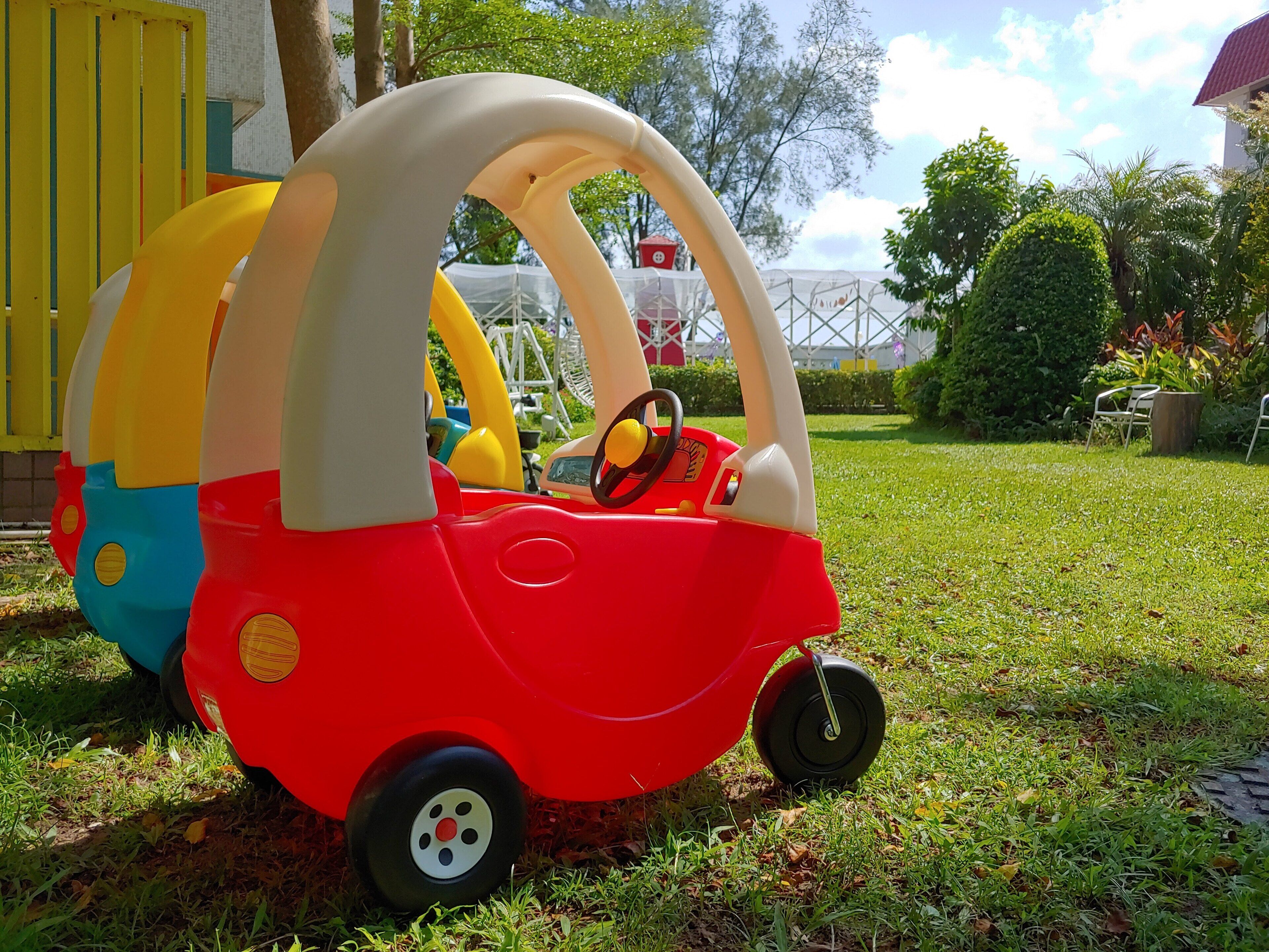 Une petite voiture rouge, avec un toit blanc et des côtés jaunes et bleus, est garée sur l’herbe.
