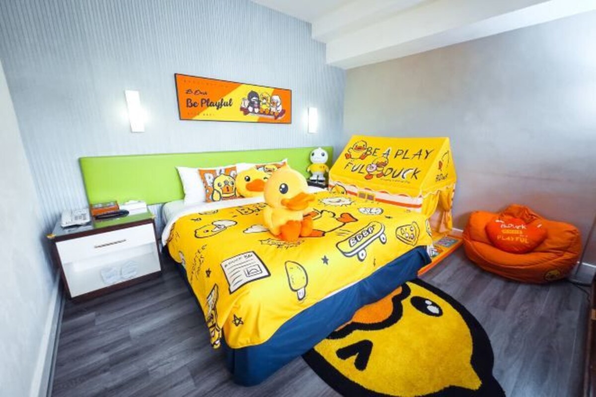 A hotel room with a bed featuring cartoon characters, a green headboard, a nightstand, and a wall with a poster.