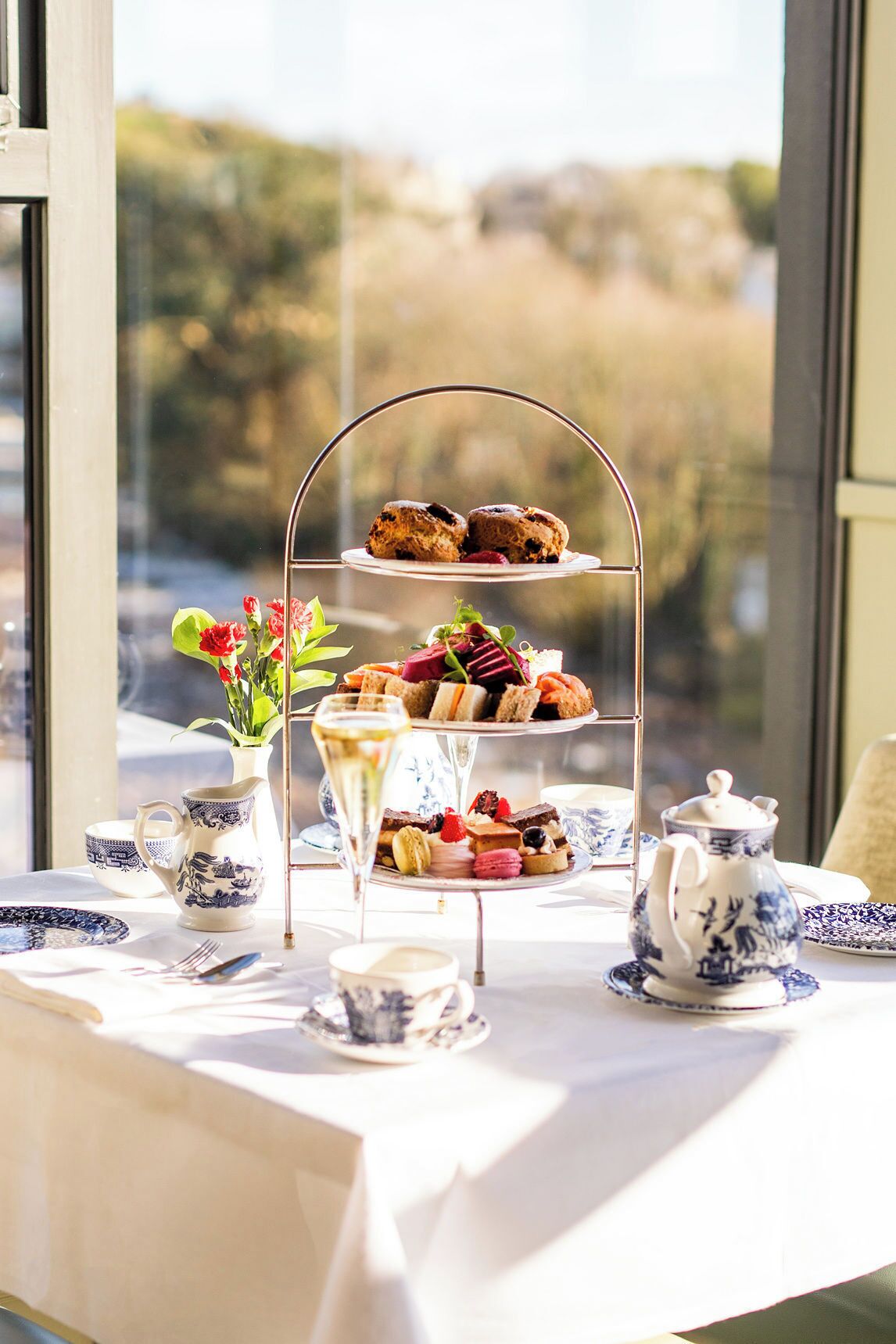 Ein mehrstufiger Dessertteller mit verschiedenen Gebäcken und einer Teekanne, platziert auf einem Tisch mit Blick auf eine natürliche Landschaft durch ein Fenster.