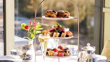 A tiered dessert stand with various pastries and a pitcher of tea, set on a table with a view of a natural landscape through a window.