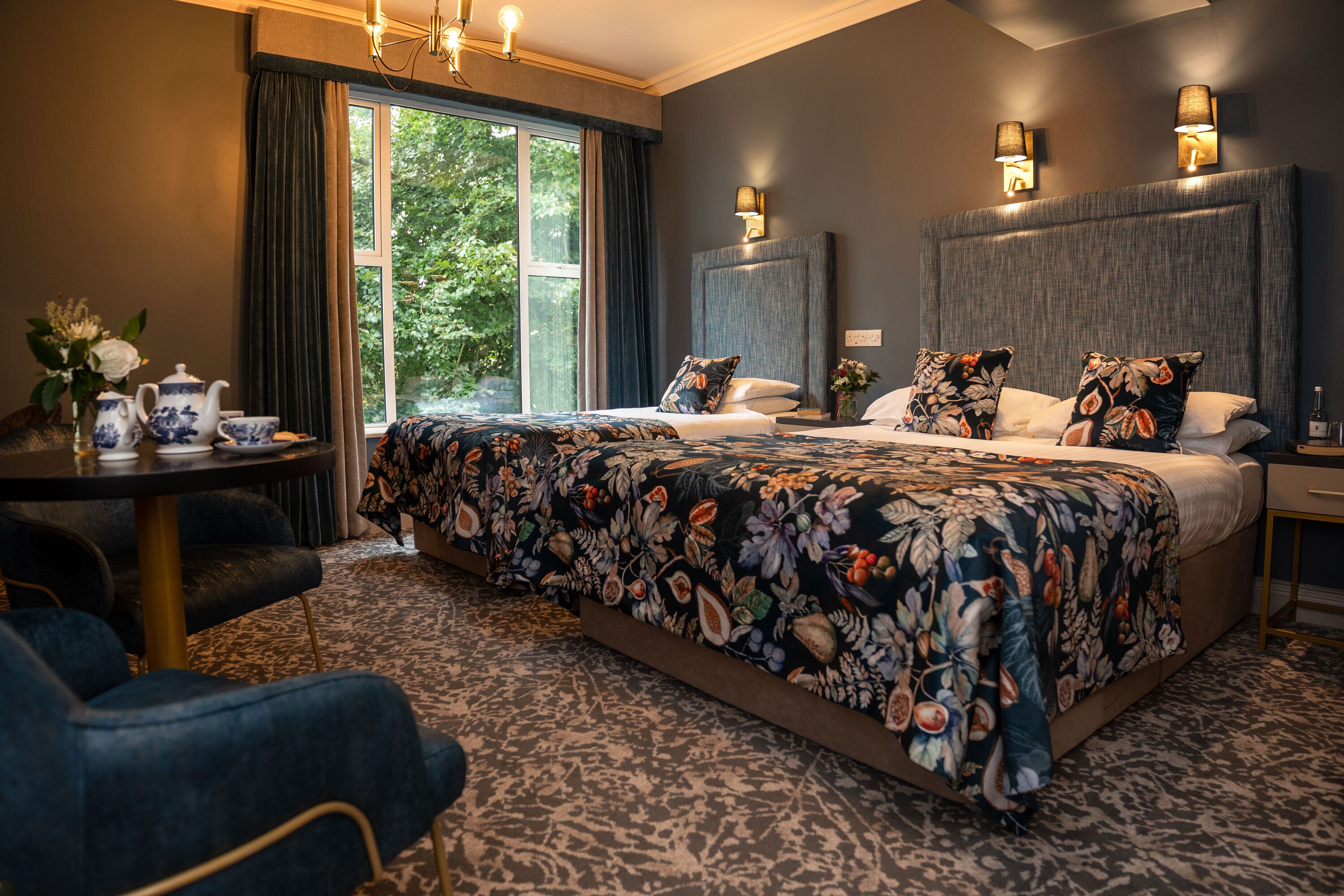 A hotel room with a large bed, a sitting area with a table and chairs, and a view of greenery outside.