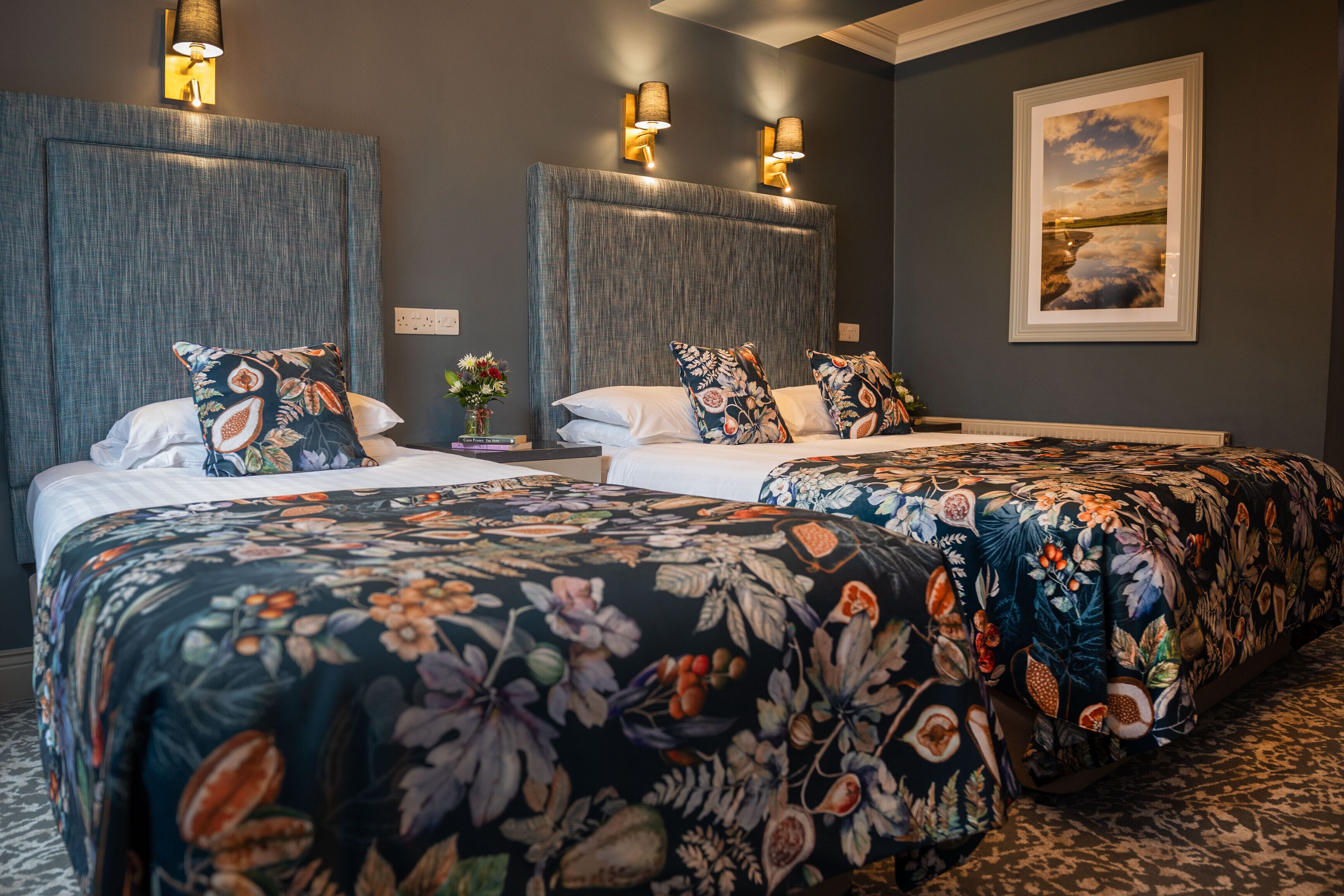 A hotel room with two beds, floral bedspreads, wall-mounted lamps, and a framed picture on the wall.