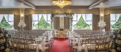 A formal event space with rows of chairs, a red carpet aisle, and a floral arch at the front.