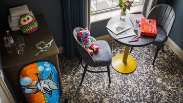 A room with a gray armchair, a small round table with a book and a red box, a colorful plush toy, a cushion with a shark design, and a window with curtains.