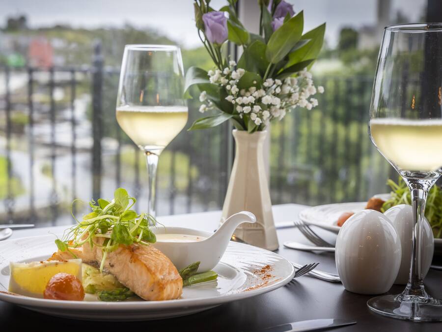 Een eettafel met een bord eten, twee glazen witte wijn en een vaas met bloemen.