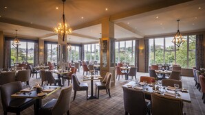 A spacious dining area with multiple tables and chairs, large windows offering a view of the outdoors, and elegant chandeliers. - Falls Hotel & Spa (Ennistimon)