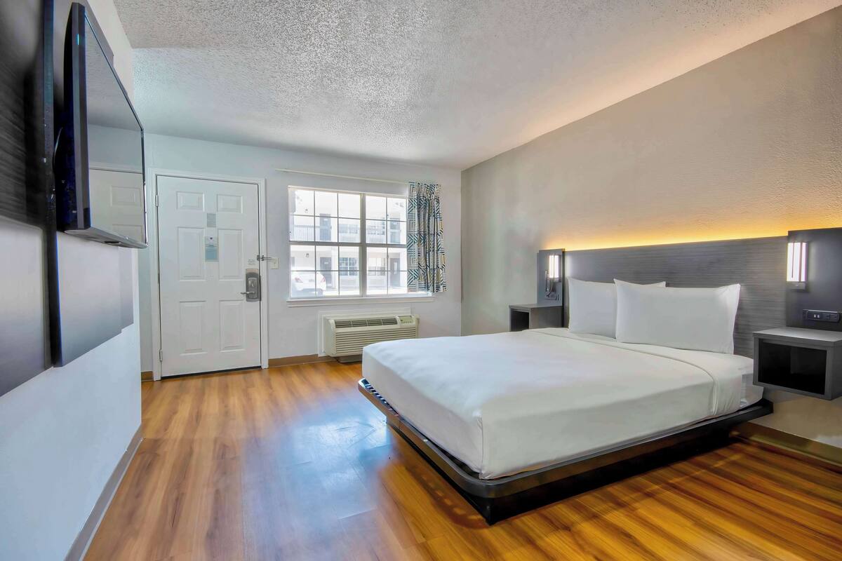 Habitación de hotel con una cama grande, un televisor de pantalla plana, una ventana con persianas, una puerta y una mesita de noche con lámpara.