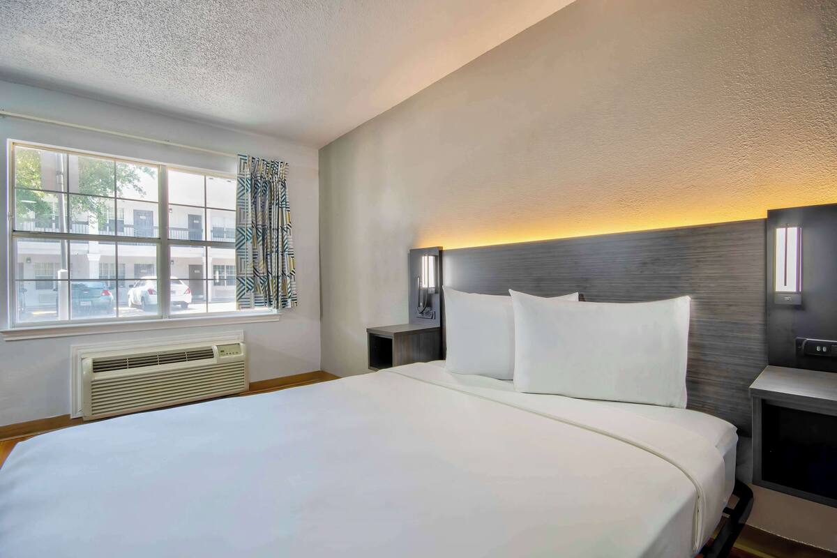 Una habitación de hotel moderna con una cama grande, una ventana con persianas, un calentador en la pared y una mesita de noche con una lámpara.