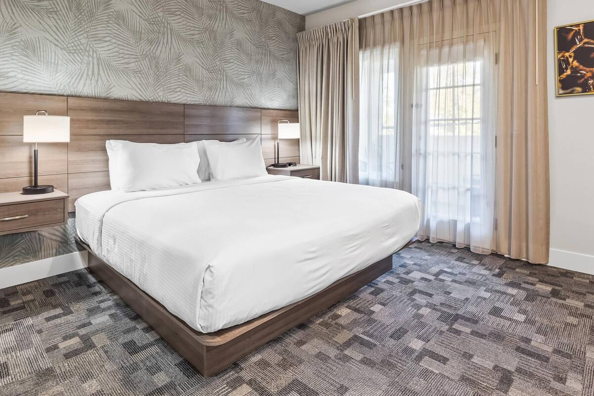Cama bem arrumada com lençóis brancos, cabeceira de madeira, duas luminárias de apoio e um tapete estampado.