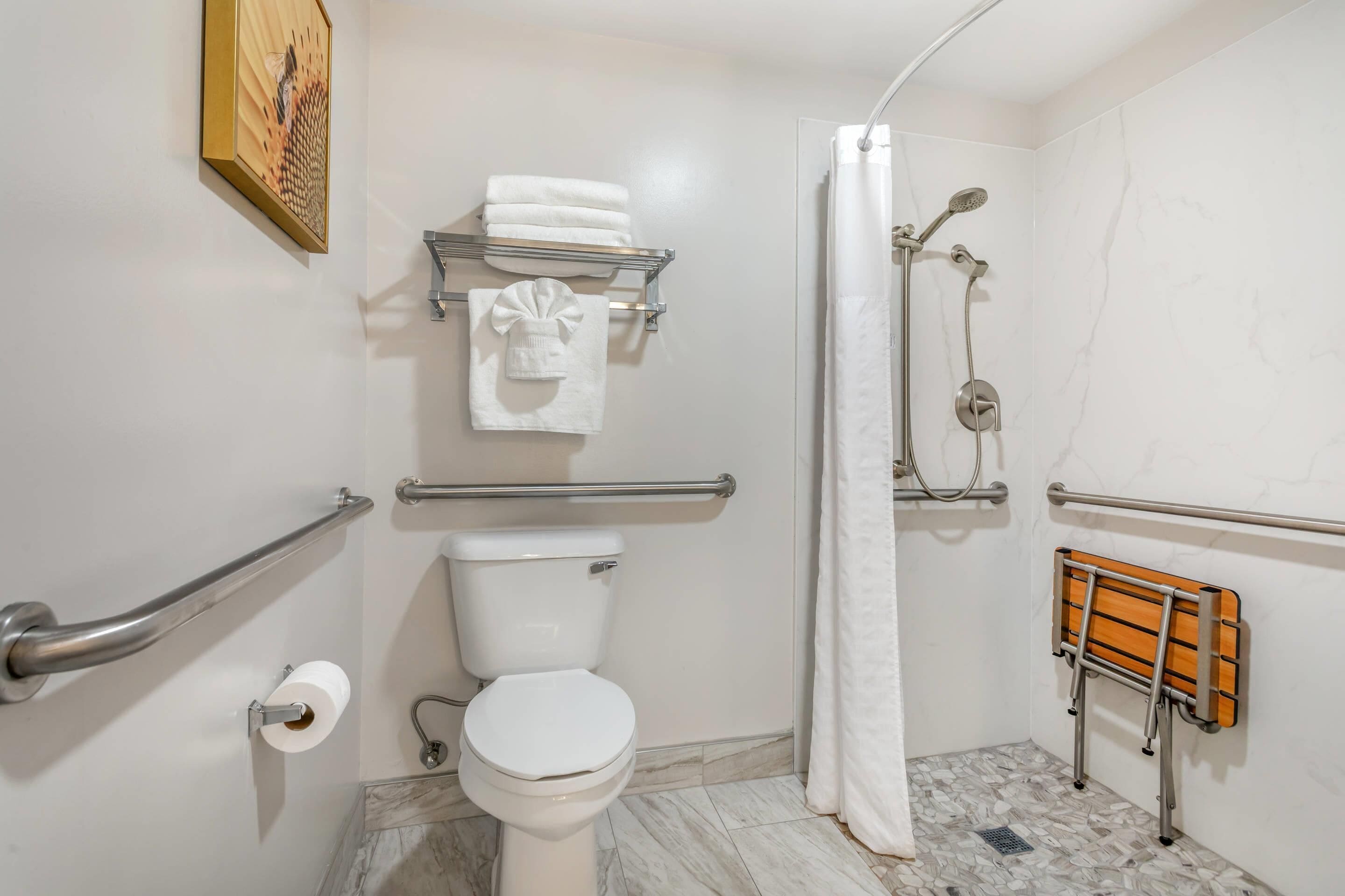 Un baño con inodoro, barras de apoyo, ducha, toallero y banco de madera.