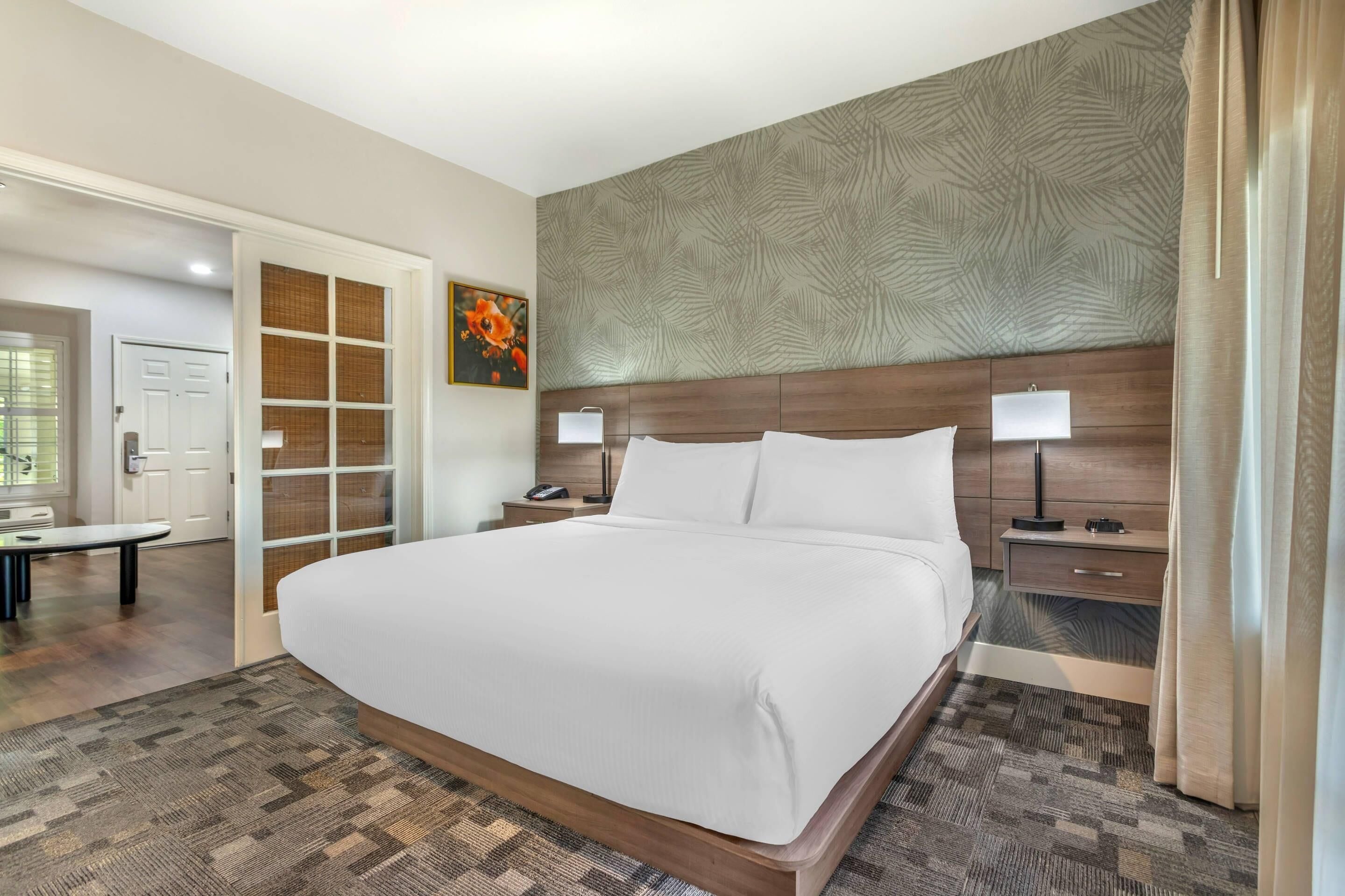 A hotel room with a large bed, wooden headboard, two bedside tables with lamps, a nightstand, a wardrobe with a mirror, and a door leading to another room.