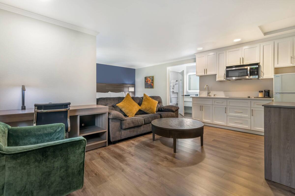 Uma sala de estar moderna com um sofá, uma poltrona verde e uma mesa de centro. Há uma cozinha com armários brancos e um micro-ondas.