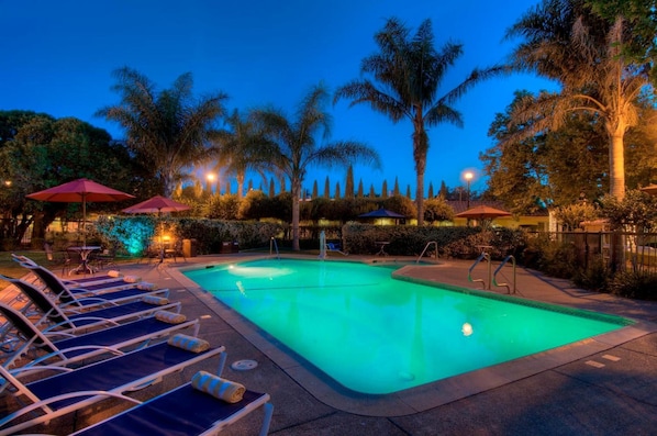 A swimming pool with lounge chairs and umbrellas, surrounded by palm trees and well-lit pathways.