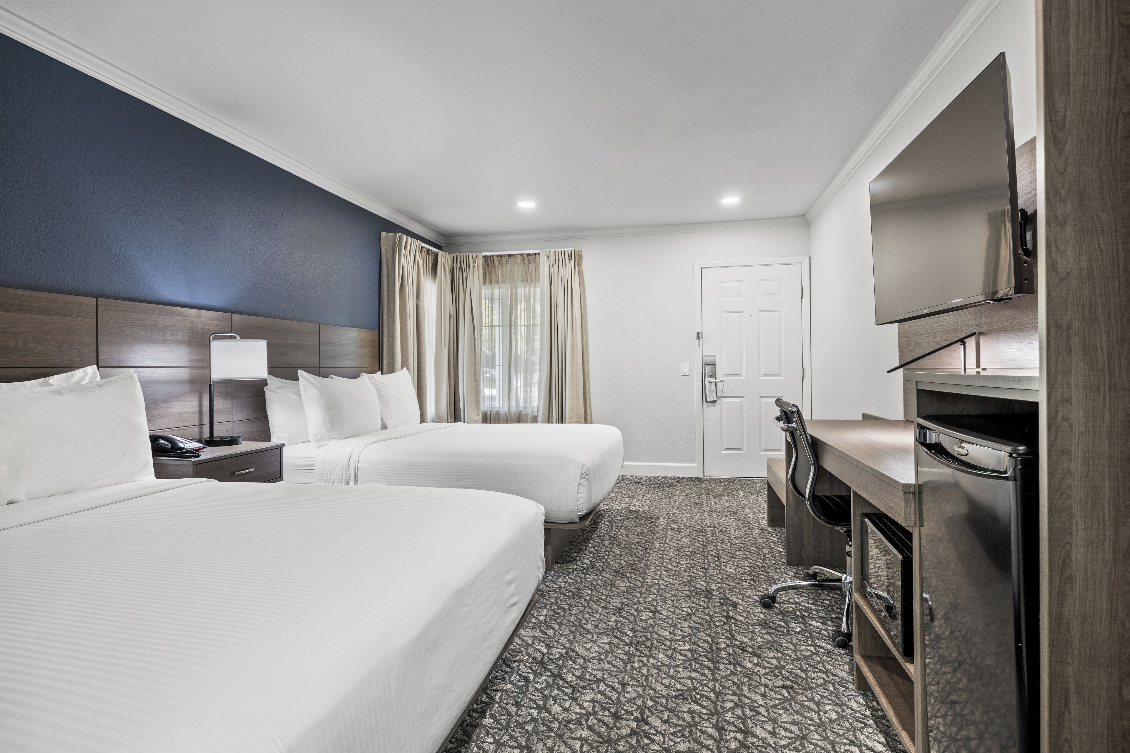 Ein Hotelzimmer mit zwei Betten, einem Schreibtisch, einem Flachbildfernseher, einer Minibar und einer Tür zum Badezimmer.