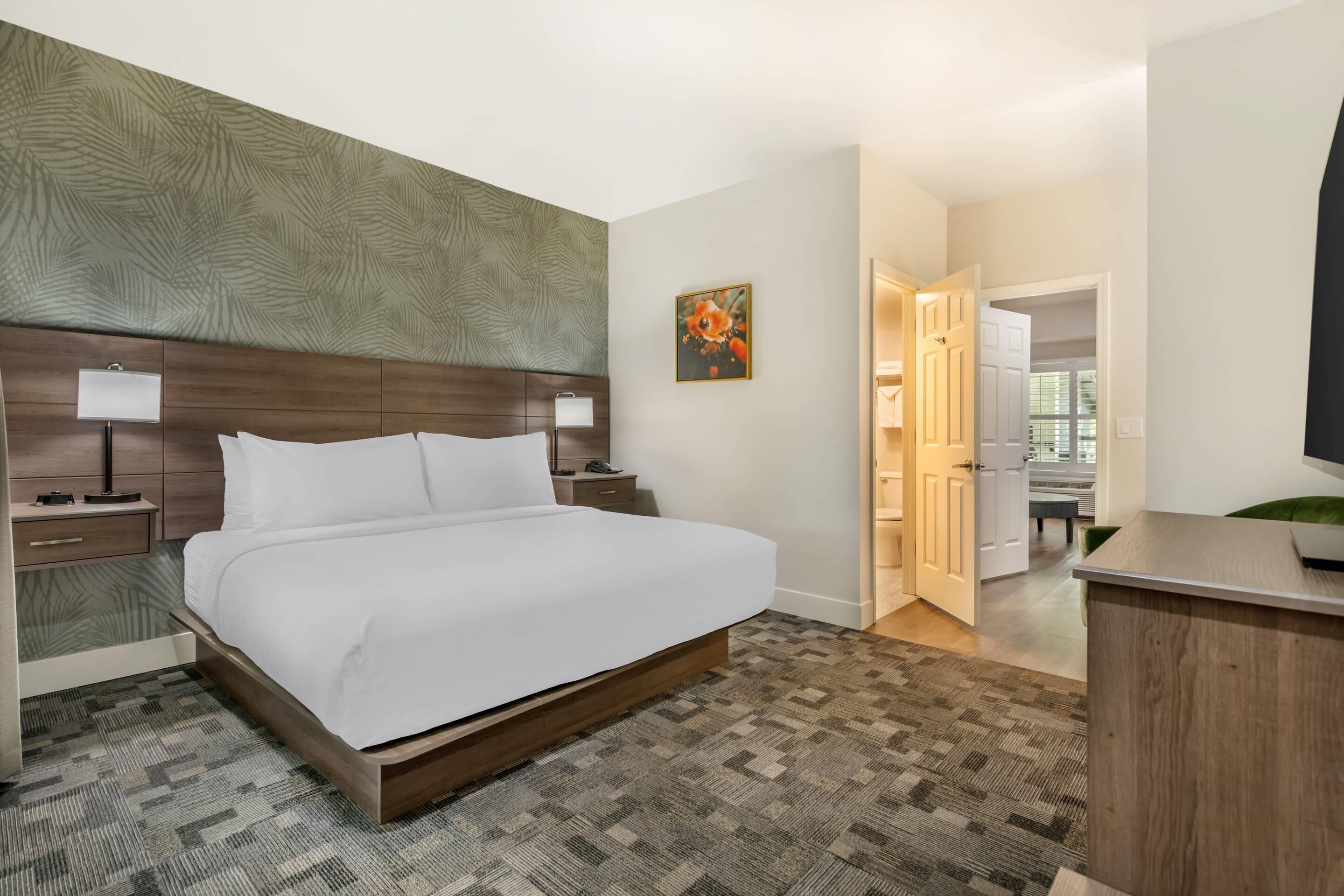 A hotel room with a large bed, a wooden headboard, a nightstand with a lamp, a painting on the wall, and a desk with a computer.