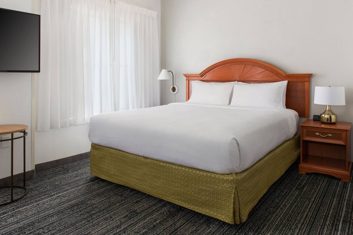 Ein Hotelzimmer mit einem großen Bett, einem Nachttisch mit Lampe, einem an der Wand montierten Fernseher und einem Fenster mit Vorhängen.