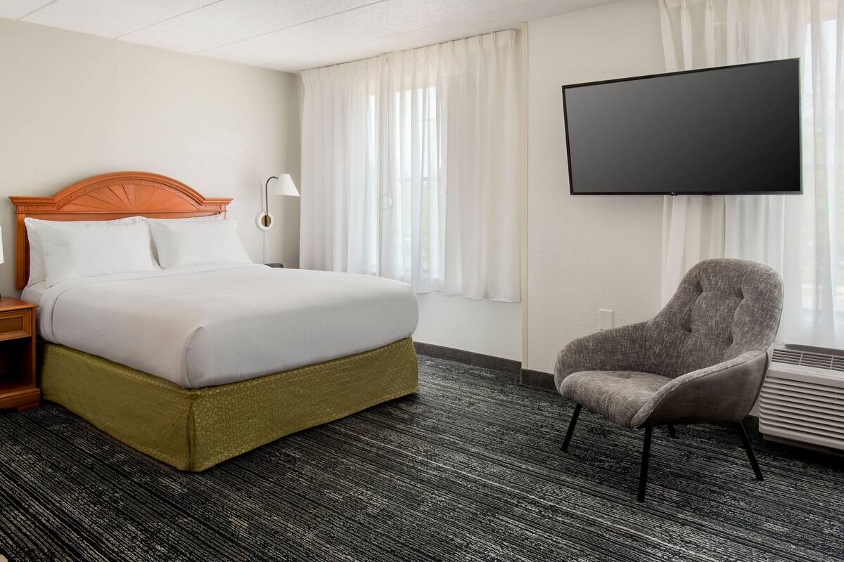 Ein Hotelzimmer mit einem großen Bett, einem Sessel, einem Flachbildfernseher und einem Fenster mit Vorhängen.