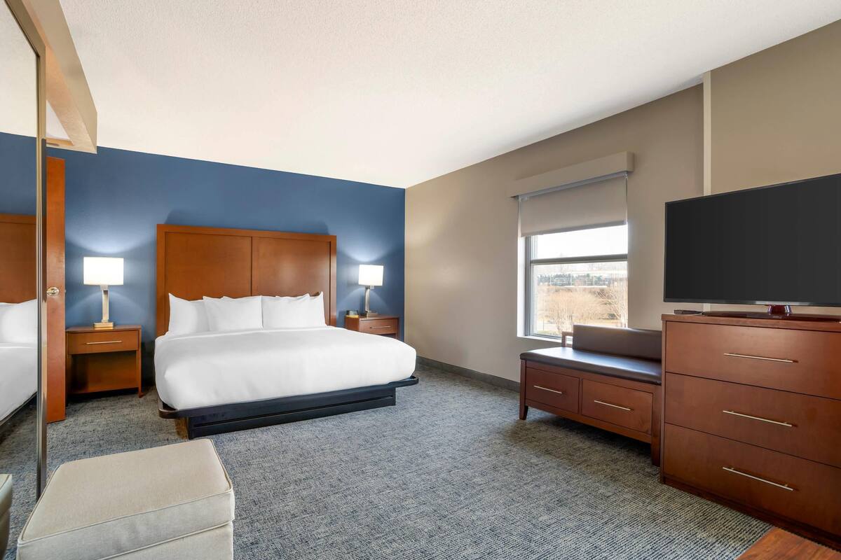 Une chambre d’hôtel avec un grand lit, deux tables de chevet avec des lampes, une commode, un téléviseur et une fenêtre avec des stores.