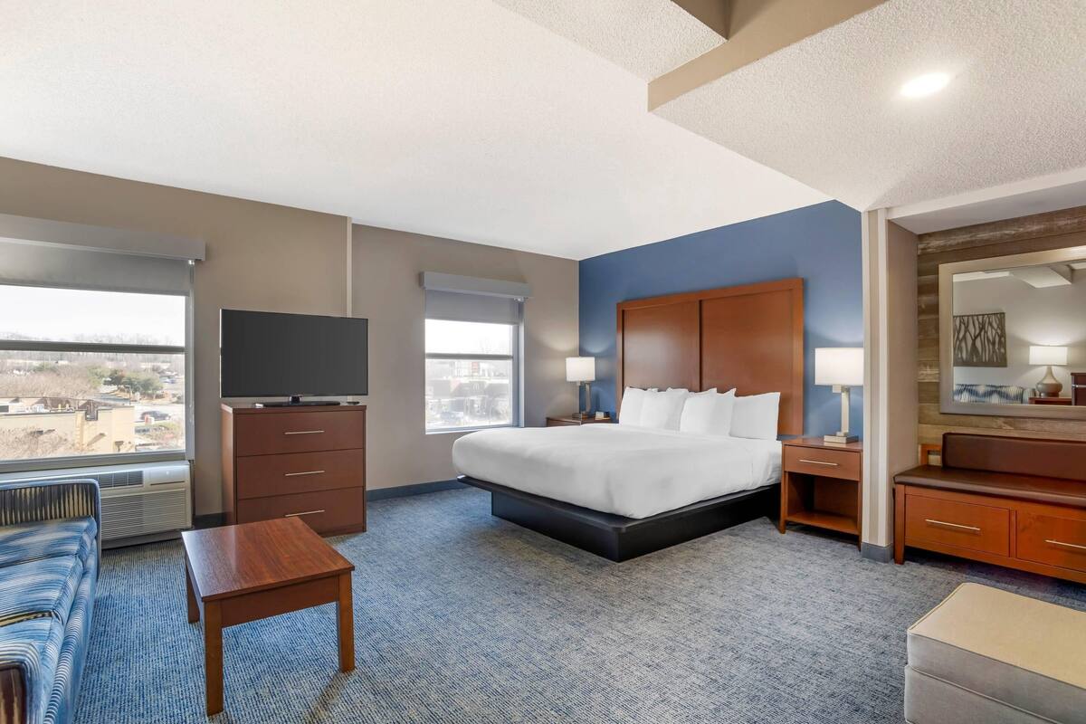 Une chambre d’hôtel équipée d’un lit, d’une télévision, d’un bureau, d’une chaise, d’une commode et d’une grande fenêtre donnant sur un paysage agréable.