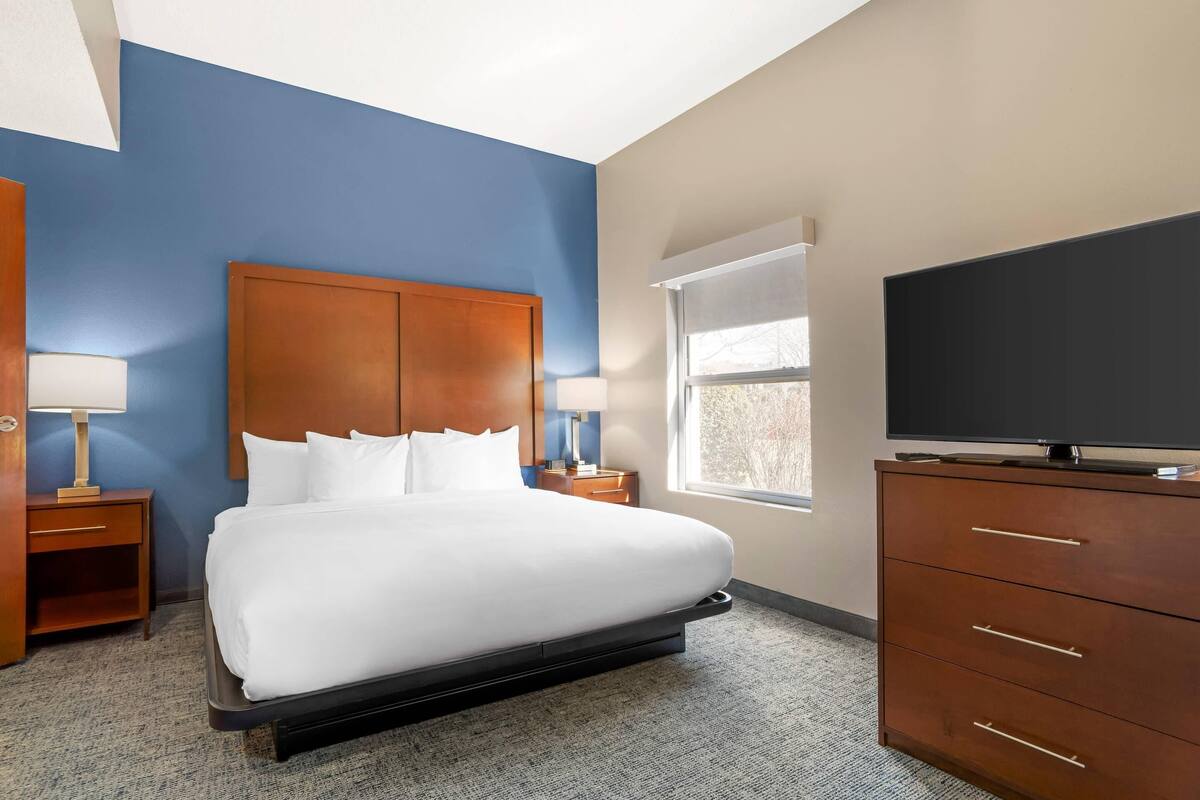 Une chambre d’hôtel avec un grand lit, une tête de lit en bois, deux tables de chevet avec des lampes, un téléviseur posé sur une commode en bois et une fenêtre avec des stores.