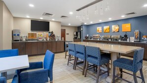 A modern dining area with a long table and blue chairs, a counter with food and beverage dispensers, and artwork on the walls. - Comfort Suites near Birkdale Village- Huntersville (Huntersville)