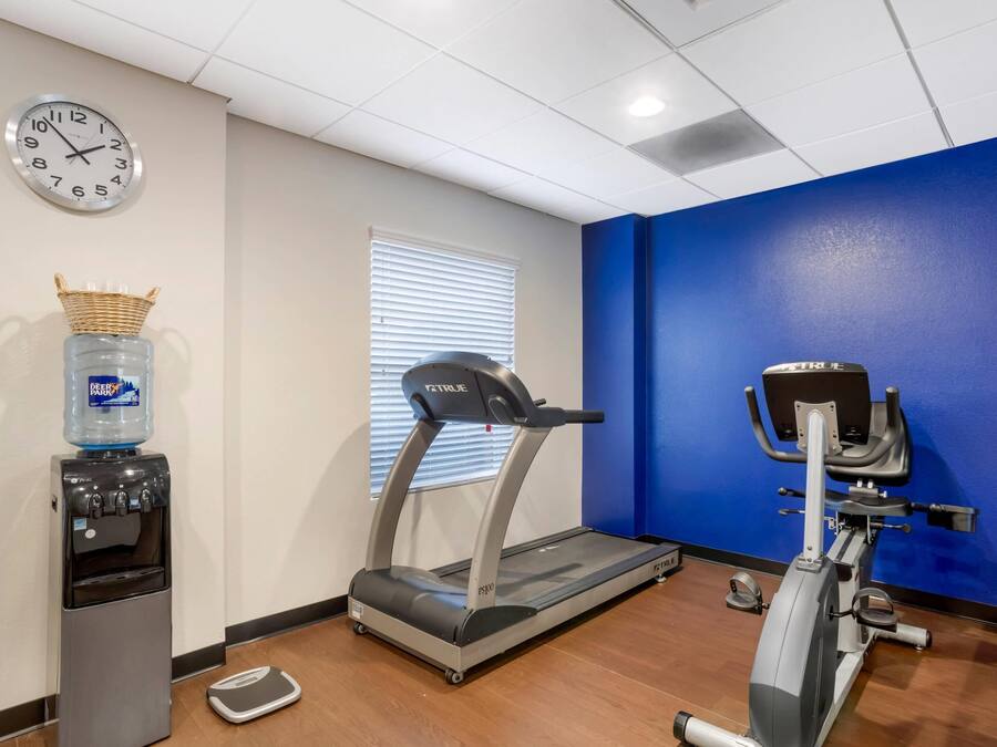 Un gimnasio con cinta de correr, elíptica, dispensador de agua, báscula y un reloj en la pared.