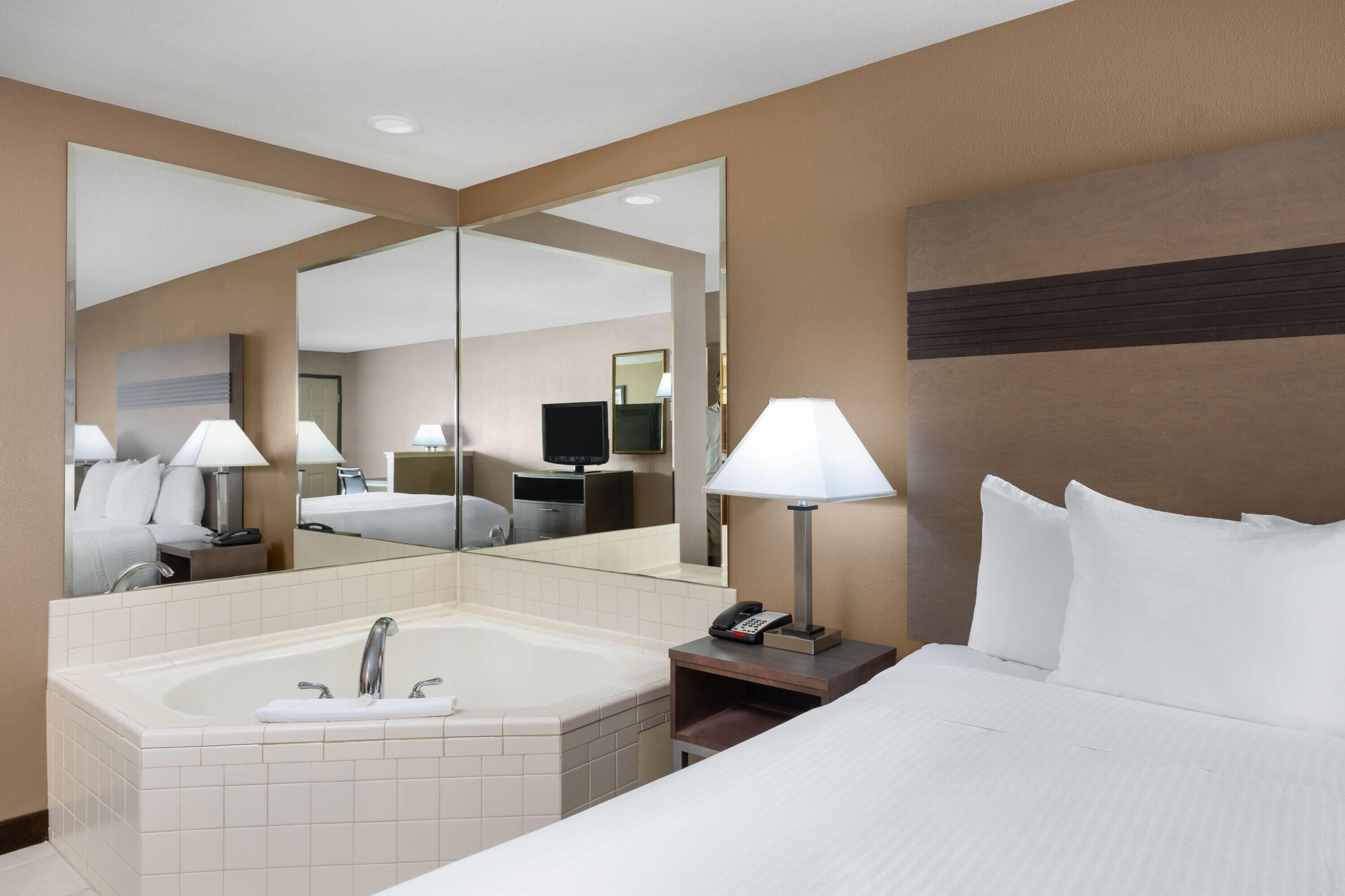 Habitación de hotel con una cama grande, baño con bañera, espejo, mesita de noche con lámpara y teléfono.