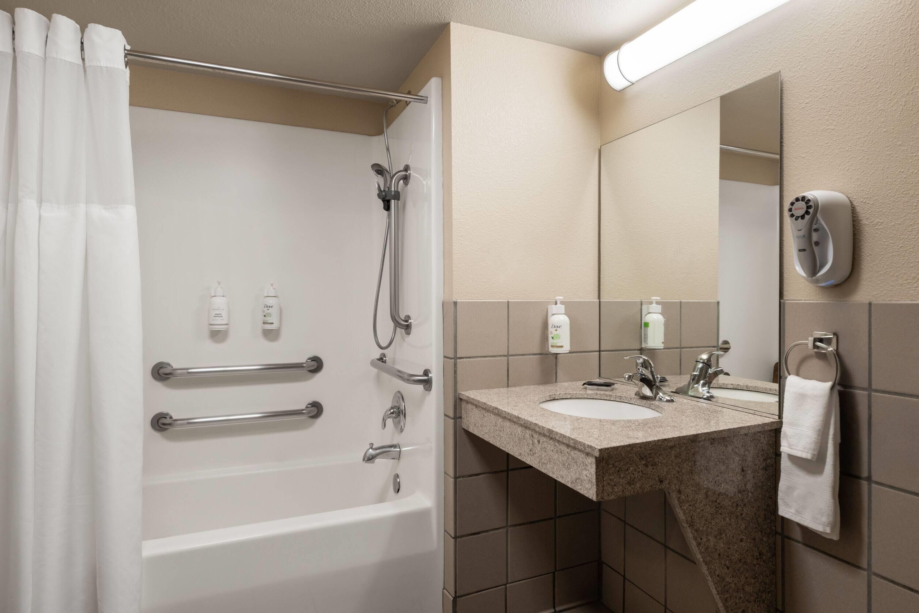 A bathroom with a bathtub, shower, and sink. There are handrails, a towel rack, and a mirror.