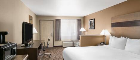Habitación de hotel con cama, escritorio con televisor, microondas, silla, lámpara de escritorio y ventana con cortinas.