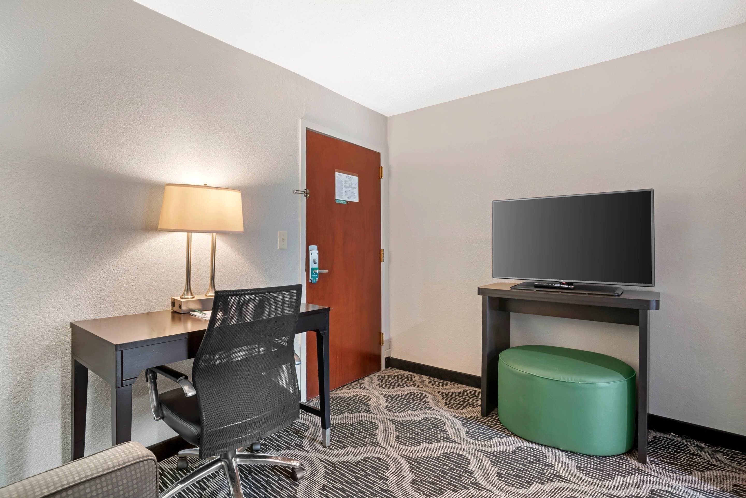 A hotel room with a desk, chair, television, and a green ottoman.