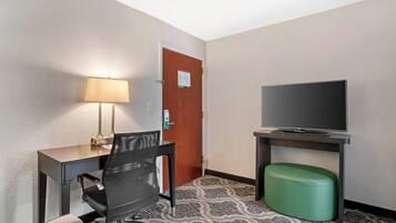 A hotel room with a desk, chair, television, and a green ottoman.