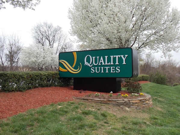 Une enseigne pour « Quality Suites », avec un logo stylisé, entourée d’un aménagement paysager soigné et d’arbres.