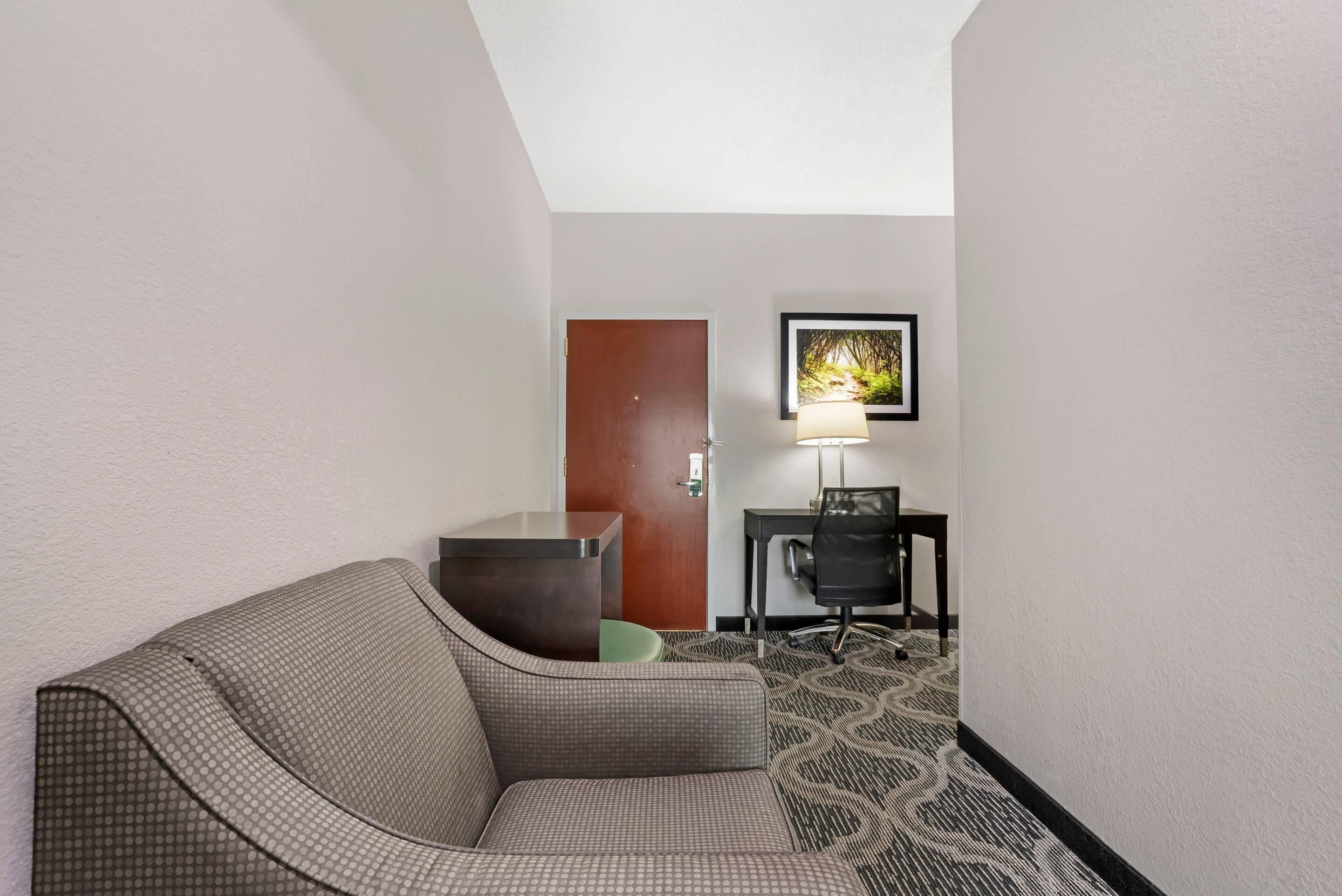 Et hotelværelse med en sofa, et skrivebord, en stol, en dør og et billede i ramme på væggen.