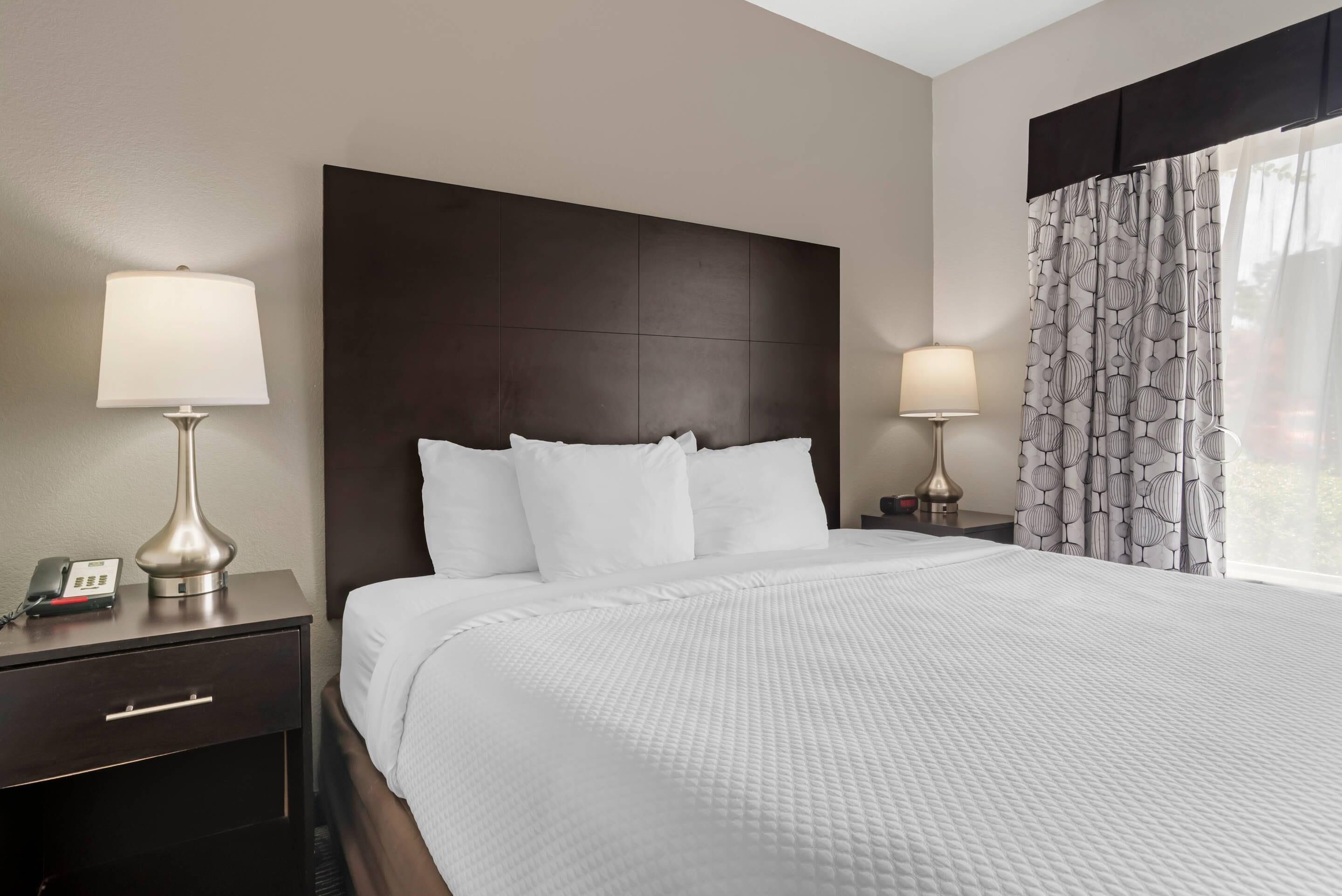 A hotel room with a large bed, two bedside tables with lamps, a telephone, and a window with patterned curtains.