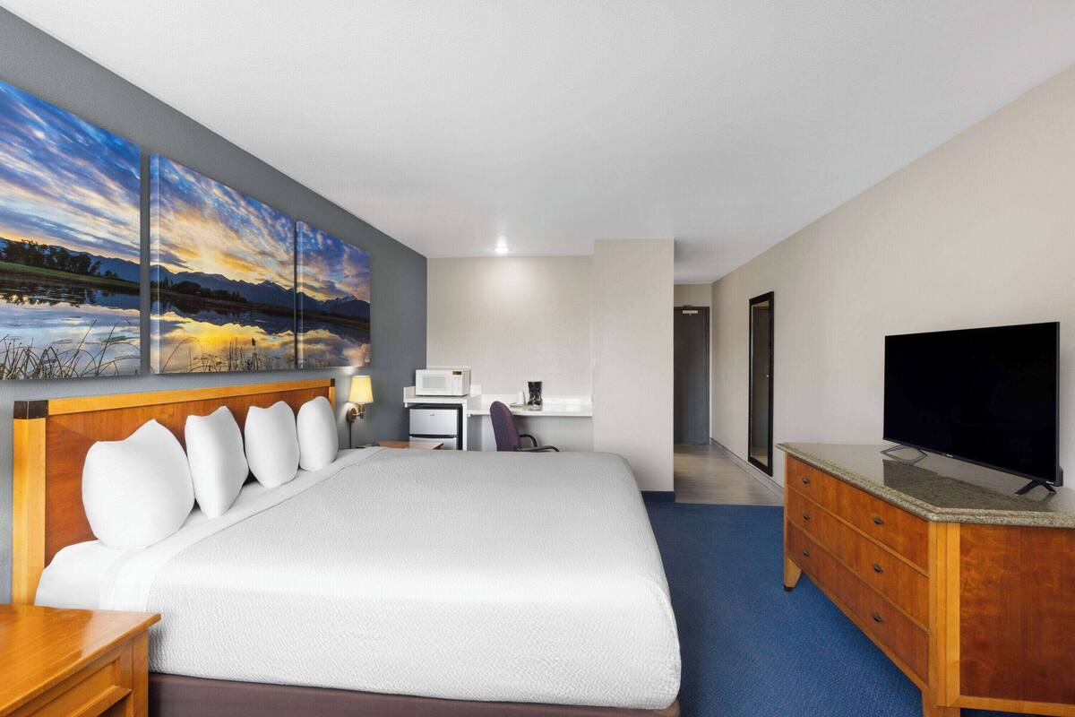 A hotel room with a large bed, a desk with a computer, a flat-screen TV, and a painting of a sunset over a lake.