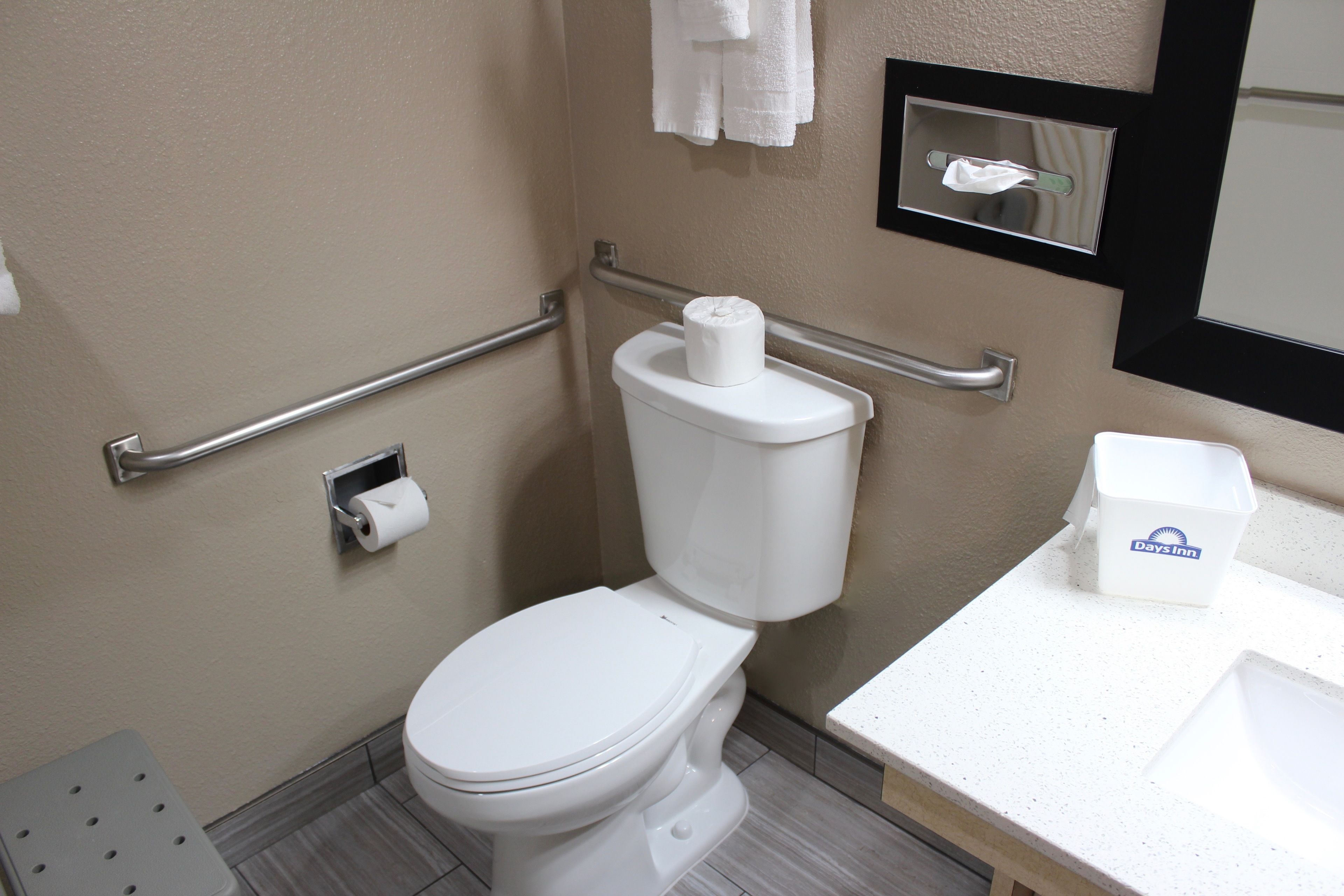 A bathroom with a white toilet, a handrail, a towel dispenser, and a sink.