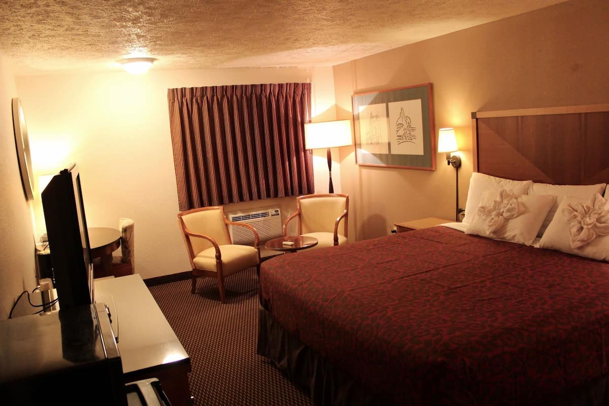 A hotel room with a bed, two chairs, a desk, a television, and a wall-mounted lamp.