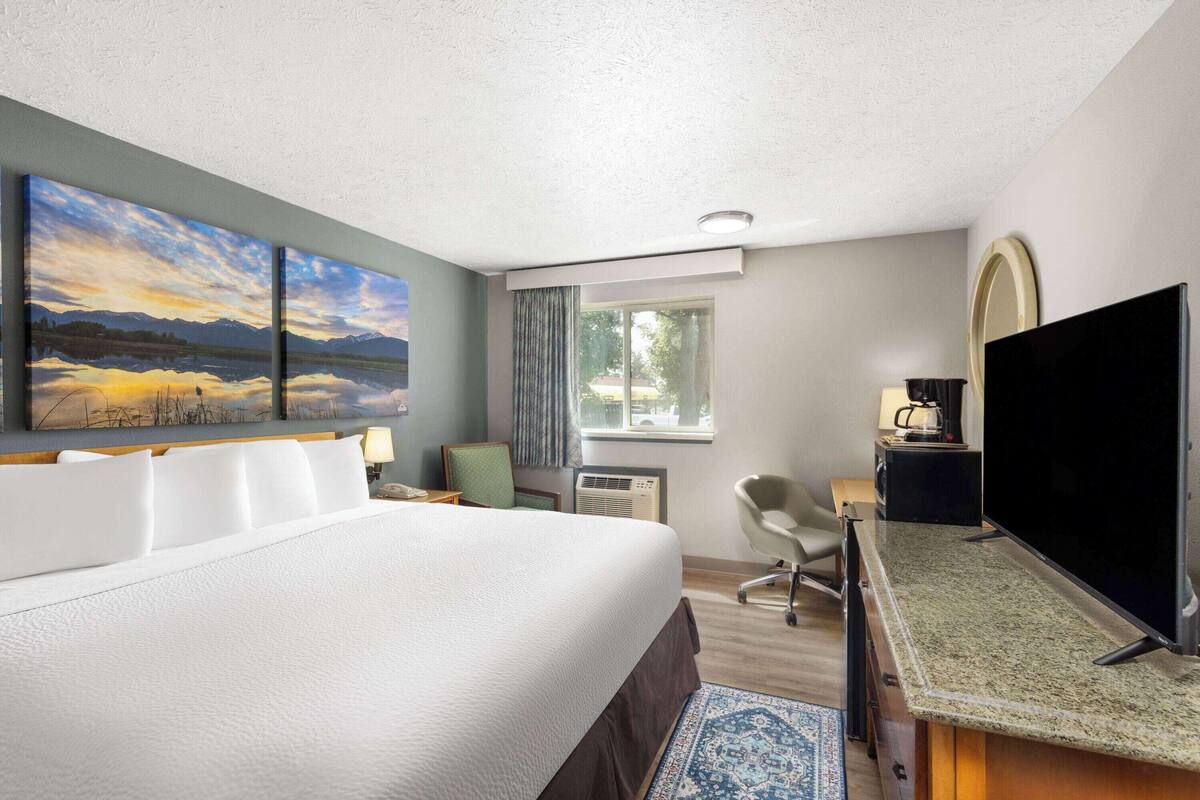 A hotel room with a large bed, a desk with a flat-screen TV, a window with curtains, and a painting on the wall.