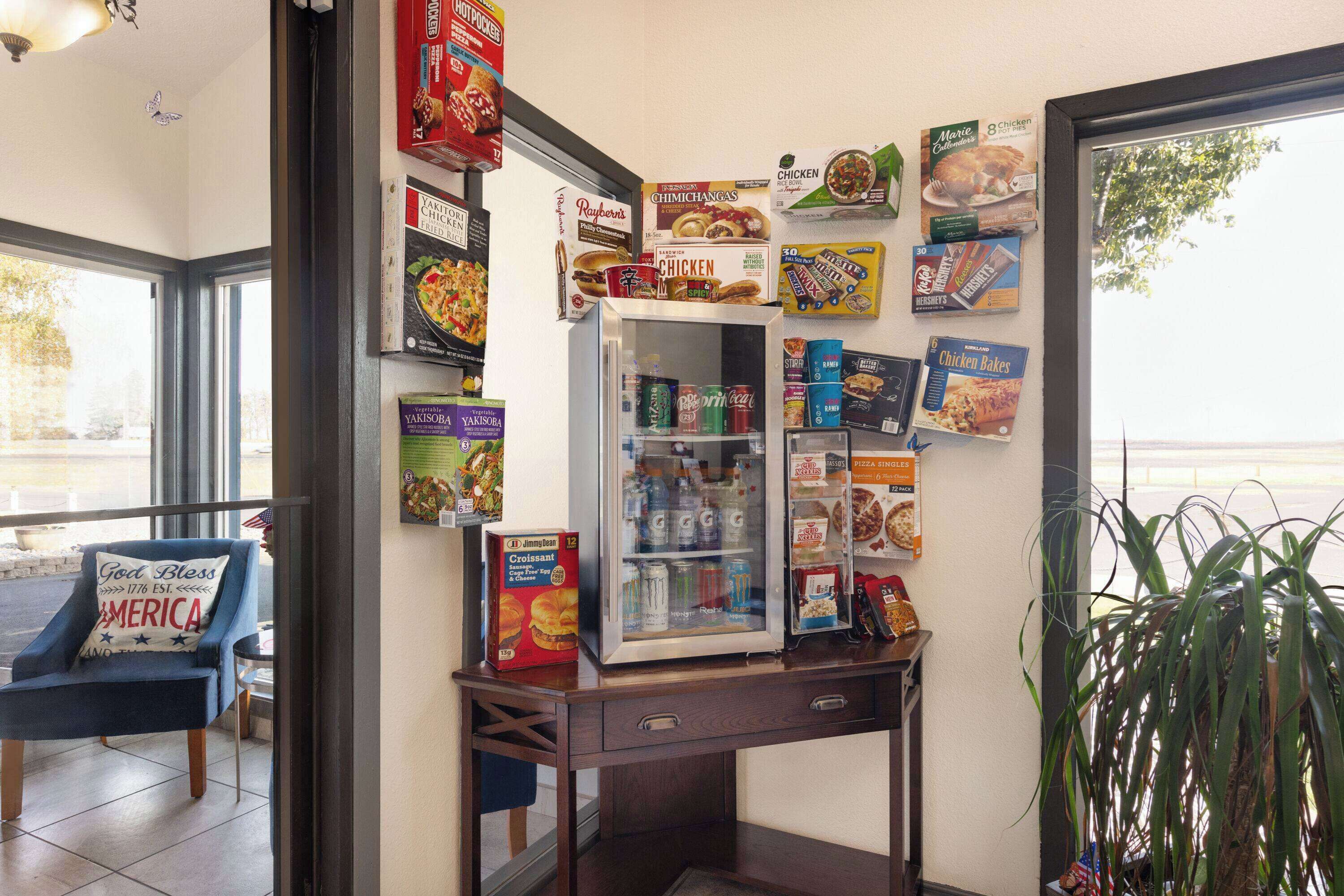A small kitchenette area with a refrigerator stocked with beverages, a microwave, and a coffee maker. There are various food and drink boxes on the wall, including brands like Chick-fil-A and Hershey's. A blue chair with a 'God Bless America' pillow is visible, and a potted plant is in the corner.