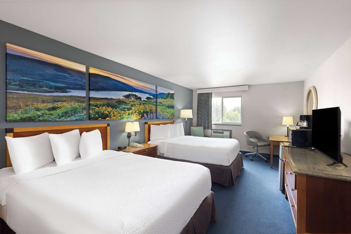 A hotel room with two beds, a desk, a TV, and a large landscape painting on the wall.