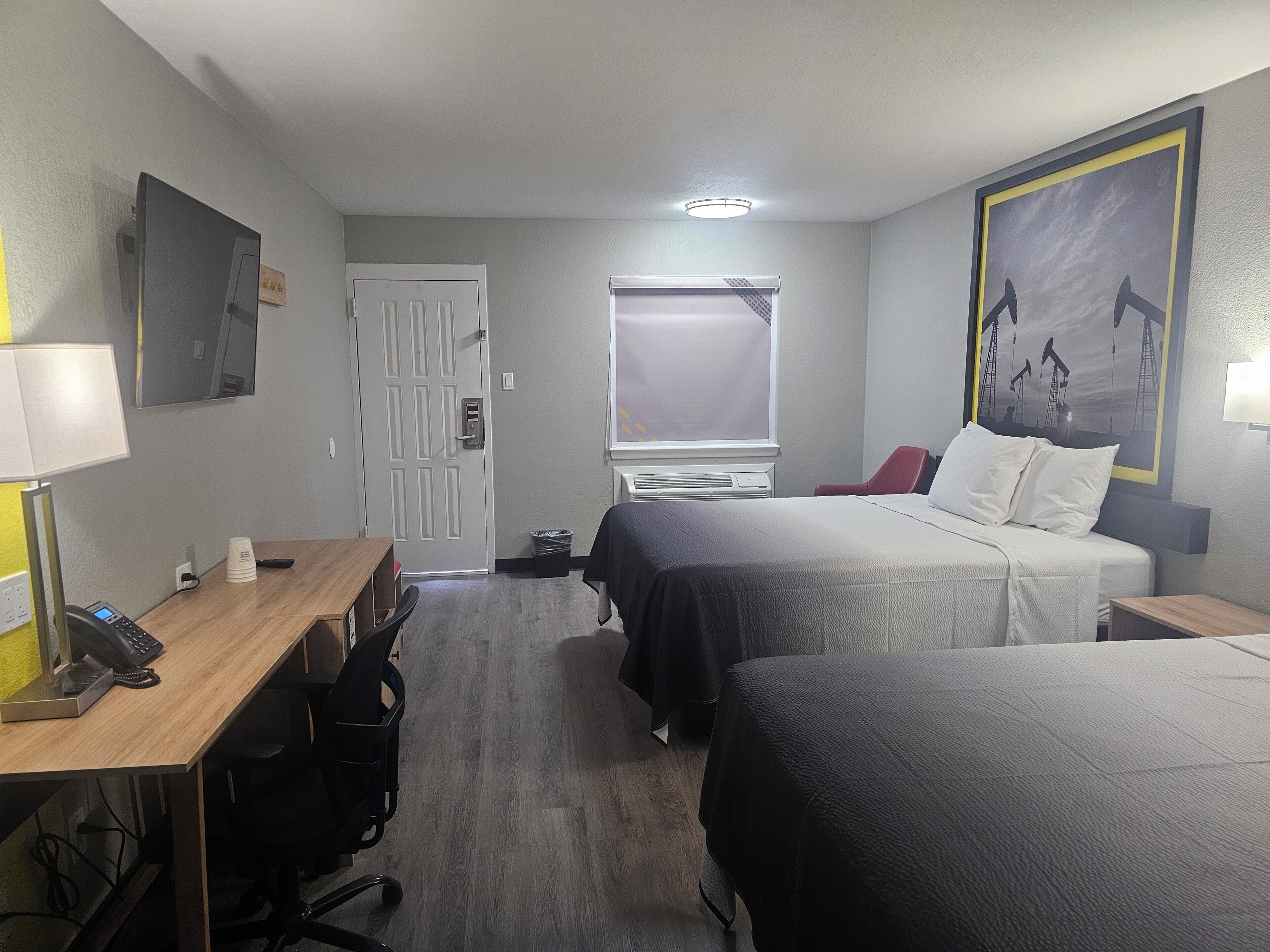 A hotel room with two beds, a desk with a phone and a lamp, a window with blinds, and a painting on the wall.